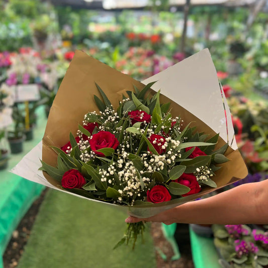 Bouquet of red roses with greenery held by a person in a greenhouse setting.