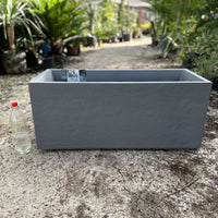 Gray rectangular planter box on a concrete surface with plants in the background