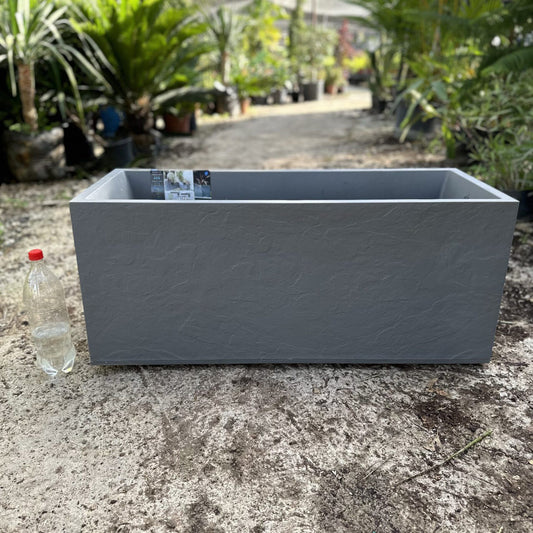 Gray rectangular planter box on a concrete surface with plants in the background